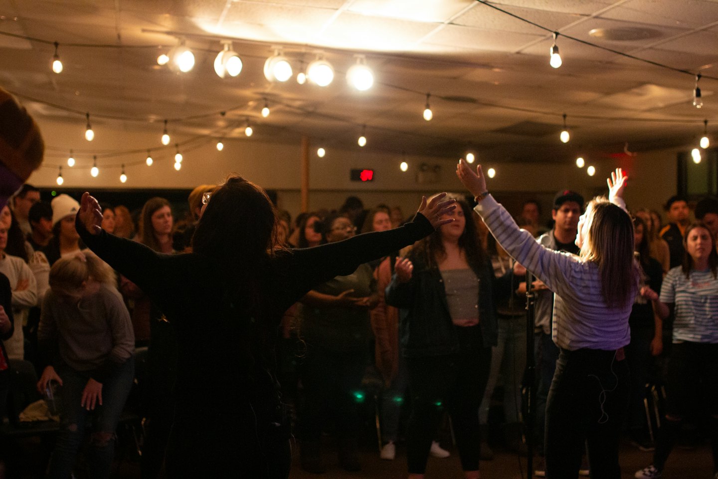 Young adults raise hands together under warm string lights.
